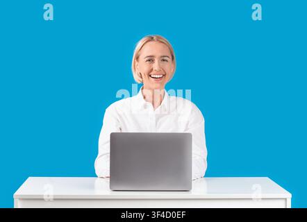 Cheerful mature business lady working with laptop at desk on blue background Stock Photo