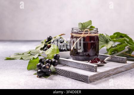 Black currant jam in a glass jar, adorned with twine, sits on a rustic wooden tray beside a bowl of fresh black currants and green leaves, with a spoo Stock Photo
