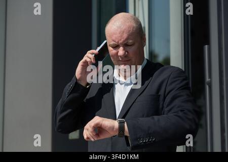 Businessman checking smartwatch while talking on smartphone outside office Stock Photo