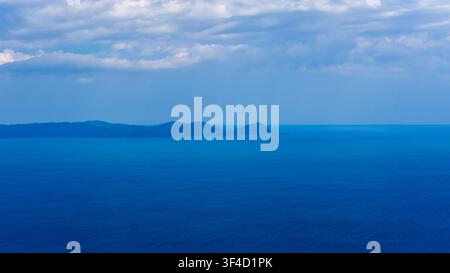 Calm deep blue water stretches toward low island silhouettes beneath layered white clouds and a soft blue summer sky in Sithonia, Greece. Stock Photo