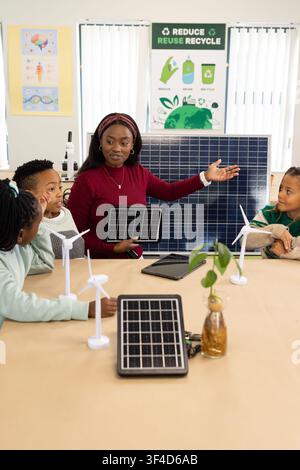 African American teacher with kids holding solar tablet gesturing toward solar panel in classroom Stock Photo