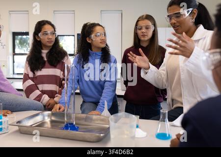 Diverse female students watching Indian instructor in lab coat at lab bench with graduated cylinder Stock Photo