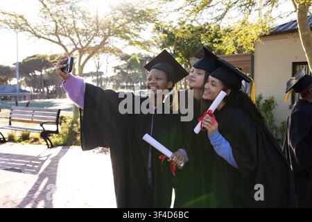 Three female graduates posing in gowns with yellow tassels holding diplomas taking selfie on campus Stock Photo