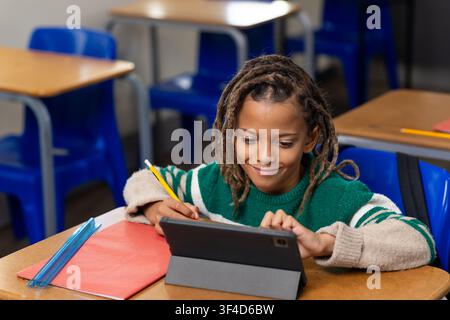 African American child sitting at school desk using tablet in folio with pencil, in green sweater Stock Photo