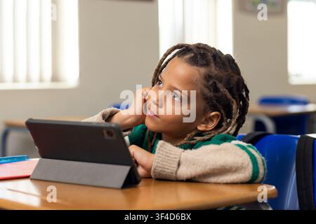 School-age African American boy sitting at desk in classroom, gazing at tablet and red notebook Stock Photo