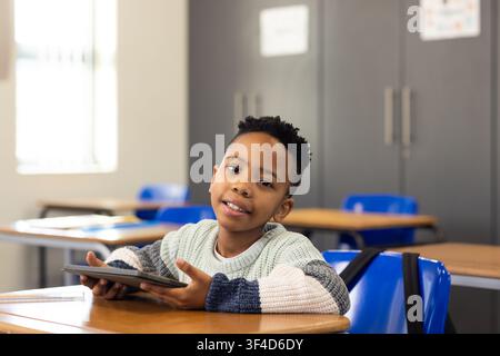 African American schoolboy holding tablet, sitting at desk in classroom with blue chairs Stock Photo