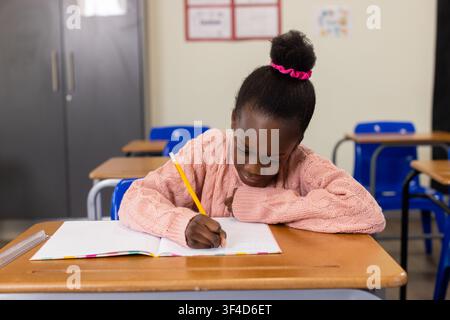 African child writing in notebook in classroom at wooden desk holding yellow pencil in pink sweater Stock Photo