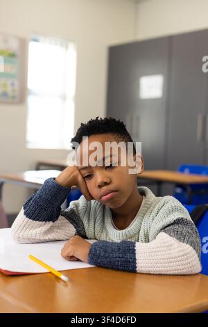 African American boy sitting at desk in classroom, knit sweater, gazing at paper with yellow pencil Stock Photo