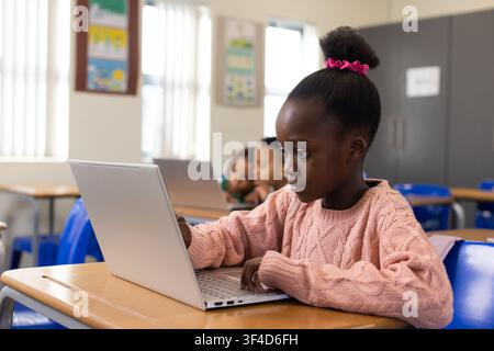African schoolgirl in pink sweater sitting at wooden desk using silver laptop at school with blinds Stock Photo