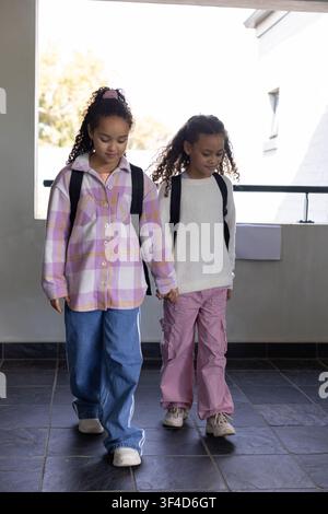 Two African American female teens holding hands walking along hall with backpacks, pink scrunchie Stock Photo