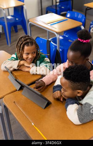 Three African American children leaning at wooden desks in classroom using two tablets Stock Photo