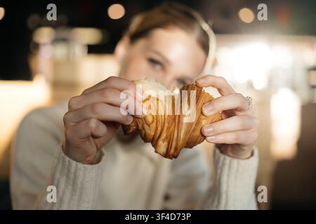 Woman holds a croissant in a cafe during daytime as she enjoys a fresh pastry Stock Photo