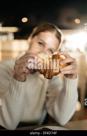 Woman holds a croissant in a cozy cafe with warm lighting during the afternoon Stock Photo