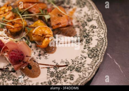A close up of a gourmet appetizer plate featuring grilled shrimp, prosciutto, raspberries, and balsamic glaze on a patterned vintage ceramic plate. Stock Photo