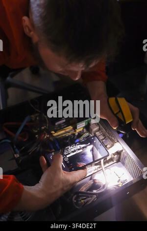 A technician in an orange shirt works on the internal hardware of a desktop computer. He holds a smartphone and a multimeter to inspect the motherboar Stock Photo