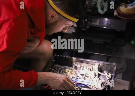 A man wearing a yellow headband and red hoodie works on a computer motherboard. He uses tools to assemble electronic components in a workshop setting. Stock Photo