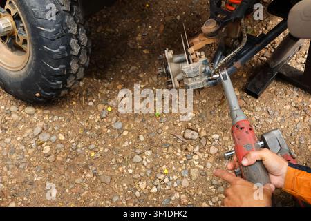 A mechanic using cordless impact wrench to remove wheel hub from off road vehicle on gravel ground, focused hands and tool in action with textured tir Stock Photo