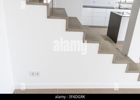 A contemporary interior features a sleek concrete staircase ascending alongside a textured white wall, leading to a modern kitchen area Stock Photo