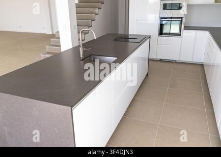 A sleek, minimalist kitchen features a dark countertop island with a stainless steel sink and induction cooktop, complemented by white cabinetry and a Stock Photo
