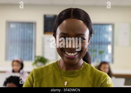 African American female teacher smiling in class in green sweater, gold pendant, students using PCs Stock Photo