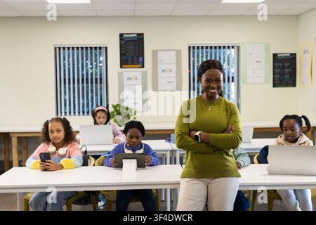 African American teacher standing front of classroom in sweater and smartwatch, children on laptops Stock Photo