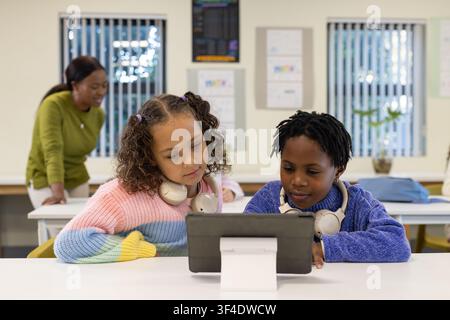 Female children sitting side-by-side at classroom table using tablet, wearing headphones, sweaters Stock Photo