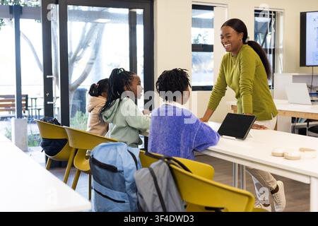 Female teacher leaning on class table engaging three female teens with tablet backpacks, copy space Stock Photo