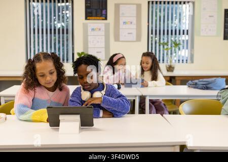 Diverse youth females sharing tablet on stand at front row in classroom, using headphones Stock Photo
