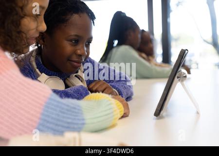 Diverse teenage female students sitting at long table in classroom, using tablet on tripod stand Stock Photo