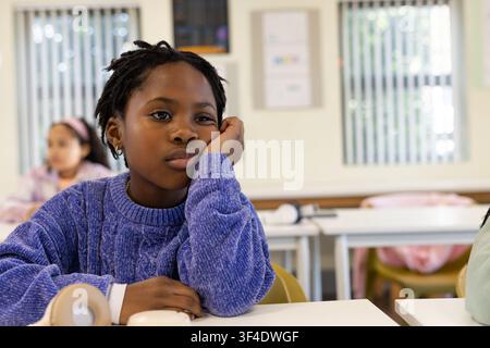 African American child sitting in classroom in purple sweater resting chin, headphones on desk Stock Photo