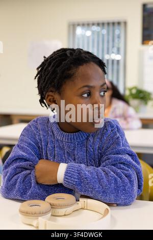 African American youth female student sitting at classroom desk wearing blue sweater beige headset Stock Photo