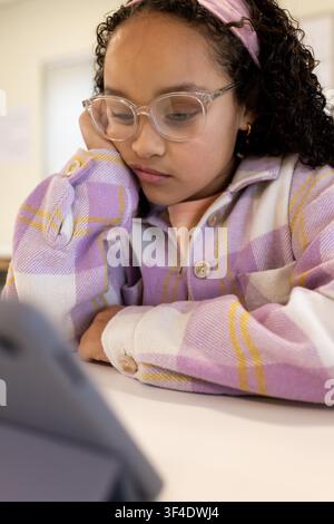 Girl child studying tablet on stand at white table in classroom wearing lilac jacket, pink headband Stock Photo