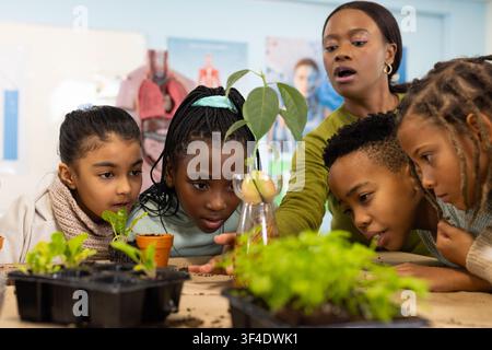 Female educator with five students leaning at school table with pots observing sprout in glass jar Stock Photo