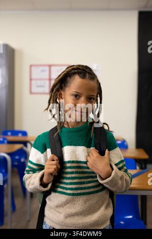 African American schoolboy standing at classroom front holding black backpack in striped sweater Stock Photo
