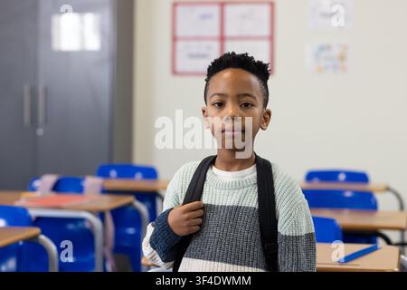 African American child standing holding backpack facing lens in class with desks blue chairs folder Stock Photo
