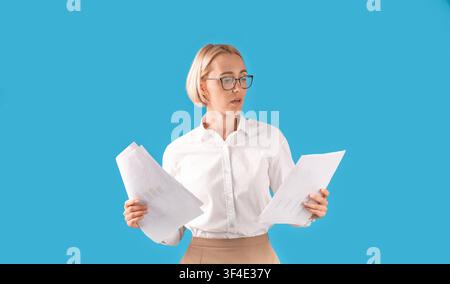 Portrait of serious businesswoman in formal clothes and eyeglasses holding documents over blue studio background Stock Photo