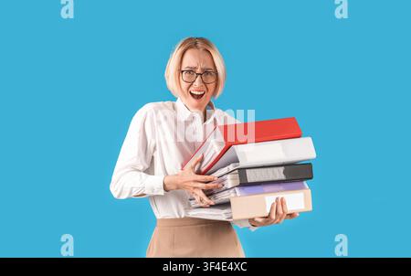 Frustrated office worker overloaded with papers, holding folders of documents on blue studio background Stock Photo