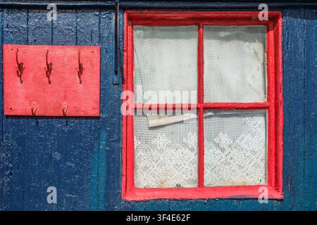A red painted window with net curtains alongside red coat hooks on and old blue wooden cabin Stock Photo