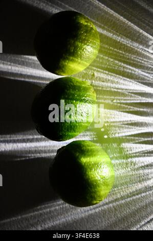 A top-down close-up of three fresh whole green limes lined up on a patterned glass tray, highlighted by strong directional lighting that creates dramatic light rays and shadow patterns. Stock Photo