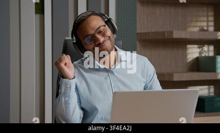 Excited happy American Indian business man dancing headphones at office desk laptop in wireless earphones having fun listening music businessman male Stock Photo