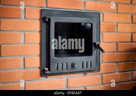 Cast iron stove door with glass built into brick wall of traditional sauna. Metal door features air vents and handle for safe wood firebox regulation Stock Photo