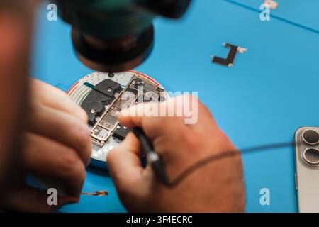 Technician repairs smartphone motherboard under magnifying glass on blue work surface with tools and components visible in the background Stock Photo