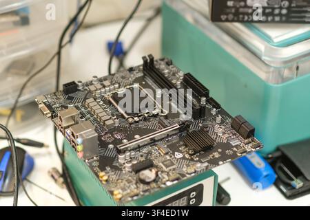 Computer motherboard placed on a workbench with various tools and equipment visible in the background of a technology workshop Stock Photo