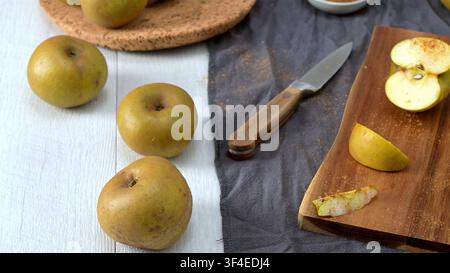 Whole and sliced golden russet apples are laid out on a rustic white surface next to a wooden cutting board, a kitchen knife, and a dark cloth under s Stock Photo