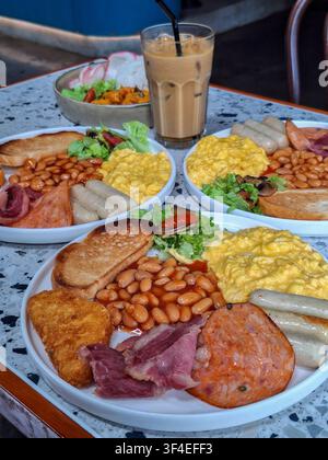 Close-up of a traditional full English breakfast plate featuring scrambled eggs, baked beans, sausages, toast, and a hash brown on a terrazzo table. Stock Photo