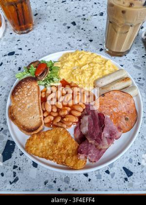 Close-up of a traditional full English breakfast plate featuring scrambled eggs, baked beans, sausages, toast, and a hash brown on a terrazzo table. Stock Photo