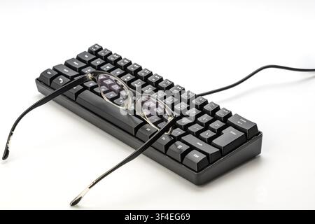 Eyeglasses on a black mechanical keyboard isolated on white background, representing nearsighted computer users, myopia, and digital eye strain Stock Photo