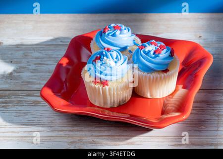 Three cupcakes with blue frosting and red stars sit on a red plate. Wooden table and blue background complete the scene. Cupcakes are in paper liners Stock Photo