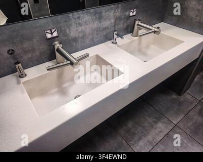 A contemporary public restroom features a long white countertop with two integrated sinks, each equipped with sleek automatic faucets and high-speed h Stock Photo