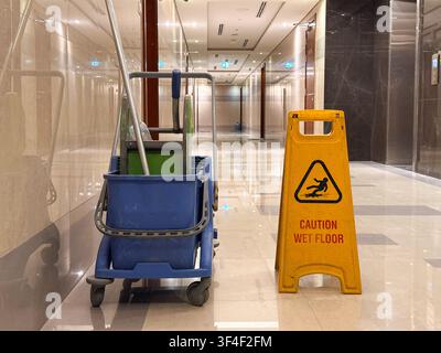 Cleaning cart with wet floor warning sign in a modern commercial hallway. Professional janitorial service, workplace safety and sanitation concept. Stock Photo